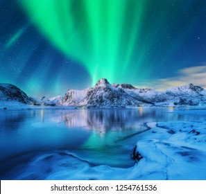 Aurora borealis über schneebedeckten Bergen, gefrorener Seeküste und Reflexion im Wasser in den Lofoten-Inseln, Norwegen. Nordlichter. Winterlandschaft mit Polarlichtern, Eis im Wasser. Sternenhimmel mit Aurora