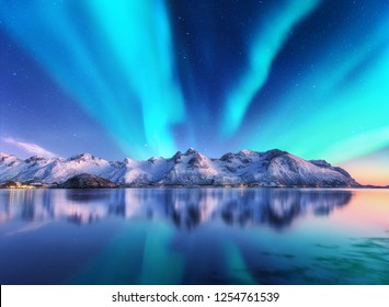 Cahaya utara dan pegunungan yang tertutup salju di pulau Lofoten, Norwegia. Aurora borealis. Langit berbintang dengan lampu kutub dan bebatuan bersalju tercermin dalam air. Pemandangan malam musim dingin dengan aurora, laut. Bepergian