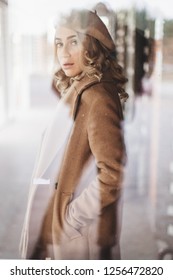 Attraktive Frau in totalem Beige-Look trägt Baskenmütze und Mantel. Lockige haare. Frau unter dem Glas des Fensters. Mädchen schaut in die Kamera. Foto im Stil des alten Films.