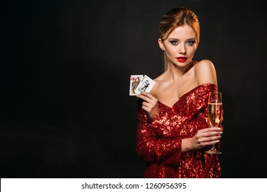 attractive girl in red shiny dress holding joker and queen of hearts cards isolated on black, looking at camera