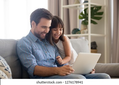 Happy father and little daughter laughing watching funny video using laptop, cheerful dad and child girl having fun playing online game on sofa together, daddy with kid relaxing at home with computer