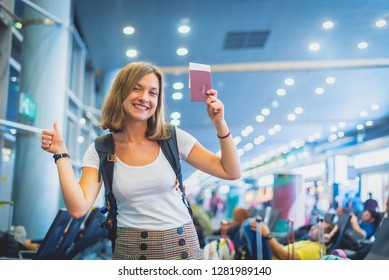 Beautiful young tourist girl in international airport, taking selfie with passport and boarding pass ready for boarding and fly. Doing gesture "ok" with other hand