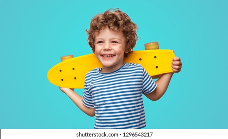 Chico rizado encantador sosteniendo pennyboard amarillo y mirando a otro lado aislado sobre fondo azul agua.