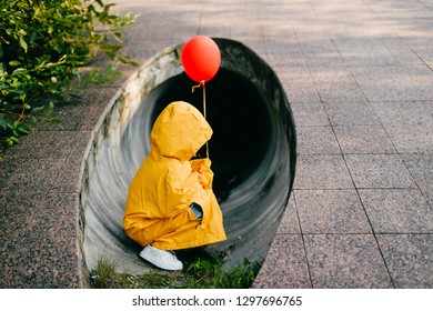 Potret gadis kecil cantik dengan jas hujan kuning besar dewasa dengan balon merah di tangan berdiri dan duduk di depan pipa terowongan limbah. Konsep film horor. Anak lucu berpose di hari musim panas