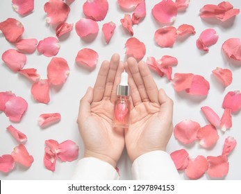 Mano femenina sosteniendo un producto casero de agua de rosas/aceite esencial para el cuidado de la piel. Pétalos de rosa rosa y frasco de vidrio cosmético con cuentagotas para suero hidratante, tónico facial, limpieza o tratamiento del acné.