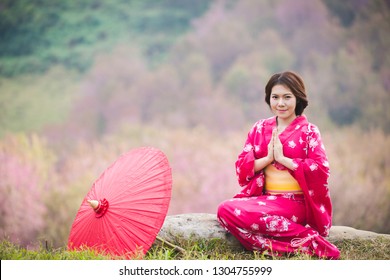 着物と赤い傘を着けたアジアの女性が、桜の花を背景にサワディーまたはタイの伝統的なハローを作っている