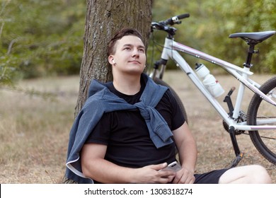 Bell'uomo caucasico con sorriso alla Monna Lisa tiene il cellulare seduto a terra vicino all'albero in un parco abbandonato. Bicicletta, auricolari, abbigliamento sportivo casual. All'aperto, look romantico, solitudine