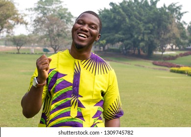 Een afrikaans mannetje in een open veld van groen gras en heldere luchten opent de hand naar de hemel in blije opluchting en lacht.