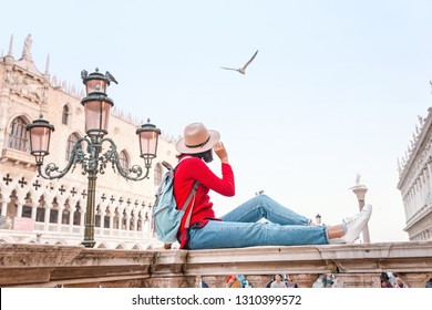Junger glücklicher asiatischer Mädchenreisender mit Hut und Rucksack, die auf einer Brüstung auf dem Markusplatz in Venedig sitzen. Urlaub und Ferien in Italien und Europa Konzept