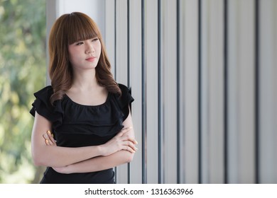 Side view portrait of young beautiful Asian lady with dark brown hair in black fashion dress standing arms crossed in modern photography studio with vertical blinds window and blur green background