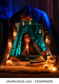 Teenage girl in the form of anime sitting in the game home tent. Scenery with fantastic garland lighting. Fantasy, dreams, divination, fairy tale, the night sky in the room.