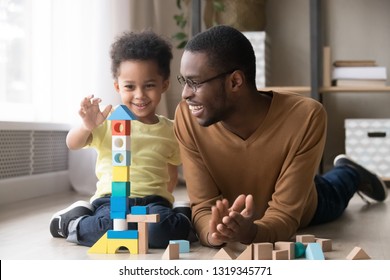 Feliz y lindo hijo jugando con papá negro niñera constructor de torre de construcción de bloques de madera multicolores, padre de familia africana y niño pequeño divirtiéndose en un piso cálido en casa