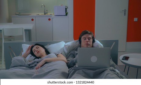 familie in bed in de ochtend. Jonge vrouw slapen. Blanke man liggend met computer en koptelefoon. Guy luistert naar muziek of kijkt naar een film in het appartement.