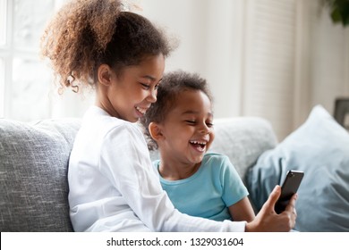 Excited small black brother and sister laugh watching funny cartoon on smartphone, smiling little mixed race siblings have fun using cellphone play game or make selfie. Kids and technology concept