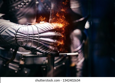 Cerca de una armadura de acero medieval con mano de guante de hierro rebosante de llamas de fuego, sosteniendo una espada gigante