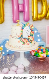 ユニコーンを使った子供の誕生日の部屋の装飾。白いレンガの壁の背景に、マシュマロ、カップケーキ、チョコレートクリーム入りブルーベリーケーキ、ガーランド、クッキーなどのお菓子が置かれたテーブルが立っています。