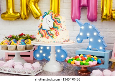 ユニコーンを使った子供の誕生日の部屋の装飾。白いレンガの壁の背景に、マシュマロ、カップケーキ、チョコレートクリーム入りブルーベリーケーキ、ガーランド、クッキーなどのお菓子が置かれたテーブルが立っています。