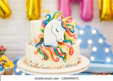 ユニコーンを使った子供の誕生日の部屋の装飾。白いレンガの壁の背景に、マシュマロ、カップケーキ、チョコレートクリーム入りブルーベリーケーキ、ガーランド、クッキーなどのお菓子が置かれたテーブルが立っています。