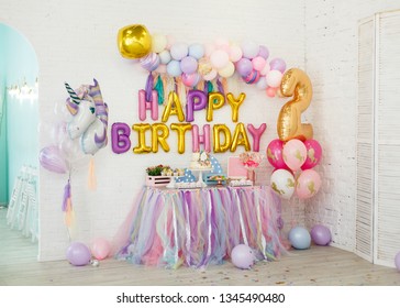 ユニコーンを使った子供の誕生日の部屋の装飾。白いレンガの壁の背景に、マシュマロ、カップケーキ、チョコレートクリーム入りブルーベリーケーキ、ガーランド、クッキーなどのお菓子が置かれたテーブルが立っています。