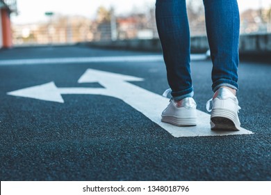 Make decision which way to go. Walking on directional sign on asphalt road. Female legs wearing jeans and white sneakers.