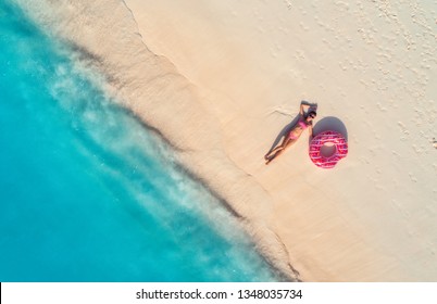 Aerial view of the beautiful young lying woman with pink donut swim ring on the white sandy beach near sea with waves at sunset. Summer holiday. Top view of slim girl, clear azure water. Indian Ocean