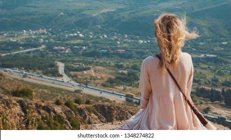 Una joven intrépida con un vestido corto de vuelo de color rosa claro se sienta en un acantilado y vigila la encantadora vista panorámica de Georgia, las maravillosas montañas cubiertas de vegetación y árboles, videos de viajeros