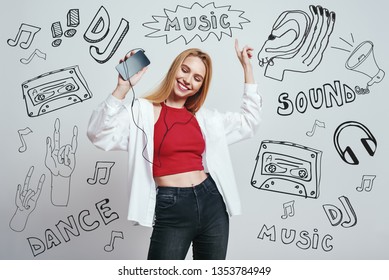 Time for music. Close up portrait of young woman in casual wear listening music with her smart phone, dancing and smiling while standing against grey background with music theme doodles