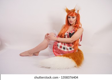 charming Fox girl in an orange long wig with fur big ears and a long fluffy tail wearing red kitchen plaid apron posing alone barefoot on a white background