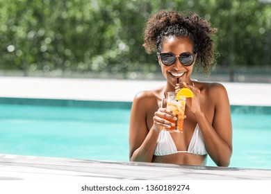 Portrait of happy fashion woman with sunglasses standing in swimming pool drinking cocktail. Beautiful african glamour girl with fresh soft drink for appetizer in luxury pool looking at camera.