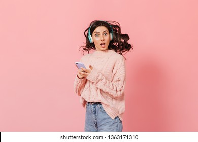 驚きの青い目をした女性は、ピンクの背景にカメラを見ます。ニットのセーターを着た女の子がヘッドフォンで音楽を聴き、iPhone を持っています
