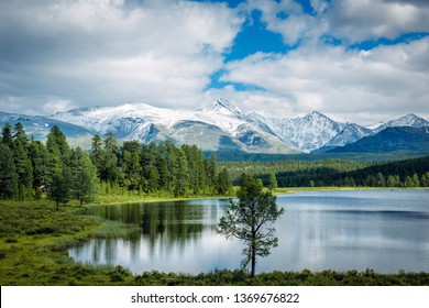 草の上の小さな湖と、緑の牧草地と雪に覆われた峰の上にふわふわの雲。ハイランド湖、アルタイ、シベリア。
