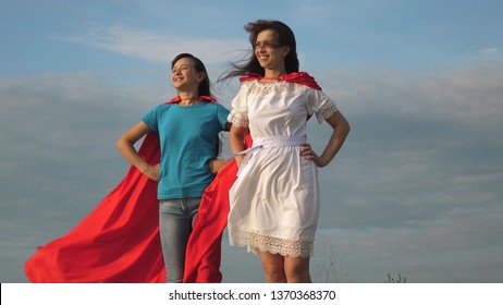 two girls in red cloaks of superheroes stand against a blue sky, wind inflates a cloak. Mom and daughter play superheroes. happy family concept