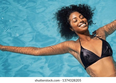 Hohe Winkelansicht des schönen Afroamerikanermädchens mit lockigem Haar und Bikini, die auf zurück in Schwimmbad schwimmen. Draufsicht der lächelnden jungen Frau sorglos im Pool mit geschlossenen Augen, die sich im Pool entspannen.