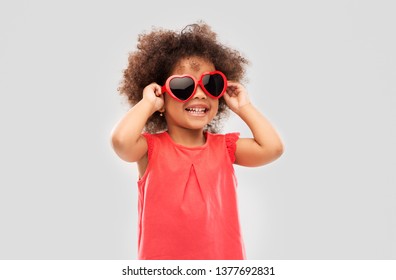 Infancia, día de San Valentín y concepto de verano - feliz niñita afroamericana con gafas de sol en forma de corazón sobre fondo gris