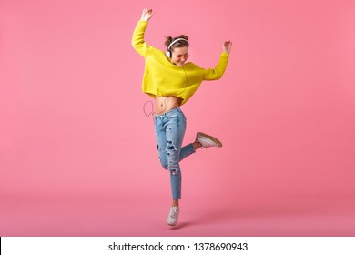 Atractiva mujer alegre y divertida saltando escuchando música con auriculares vestida con un traje colorido estilo hipster aislado sobre fondo de estudio rosa, con suéter amarillo y gafas de sol, divirtiéndose