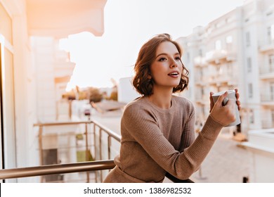 Garota sonhadora de olhos escuros tomando chá na sacada. Foto de modelo feminina caucasiana bem vestida segurando uma xícara de café.