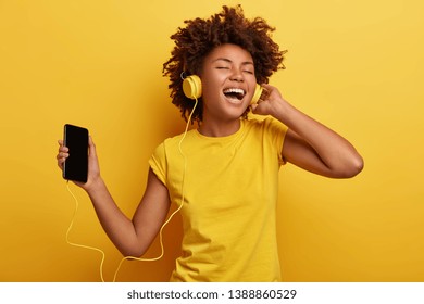 Khái niệm con người, âm nhạc, cảm xúc. Bạn nữ vô tư thỏa thích với kiểu tóc Afro nhảy múa theo nhịp điệu của giai điệu, nhắm mắt nghe bài hát lớn trong tai nghe, cầm smart phone thật vui. Màu vàng chiếm ưu thế