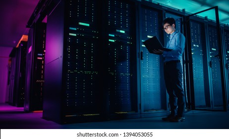 In Data Center: Male IT Technician Running Maintenance Programme on a Laptop, Controls Operational Server Rack Optimal Functioning. Modern High-Tech Operational Super Computer in Neon Colours, Lights