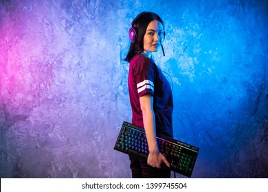 Beautiful Friendly Pro Gamer Streamer Girl Posing With a Keyboard in Her Hands, Wearing Glasses. Attractive Geek Girl with Cool Neon Retro Colors in Background.