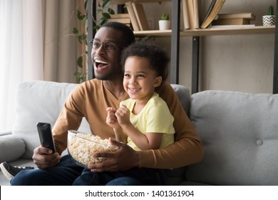 Happy African American father with toddler son, family watching tv, cartoons or football match, eating popcorn snack, sitting on couch together, little boy sitting on smiling dad knees at home