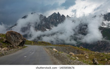 Leere asphaltierte Straße im ländlichen Frankreich führt an den felsigen Bergen vorbei, die im Morgennebel bedeckt sind. Weiße Wolken umhüllen die spektakuläre Bergkette, die sich über der malerischen Route des Grandes Alpes erhebt.