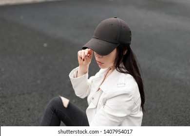 Pretty urban young woman in fashionable black baseball cap in a stylish white leather jacket relaxes and looks down outdoors. Modern American girl model sitting on the road. Trendy youth clothing.
