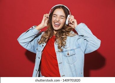 Chân dung một phụ nữ trẻ sành điệu vui vẻ mặc áo khoác denim đứng cách biệt trên nền đỏ, nghe nhạc bằng tai nghe và khiêu vũ
