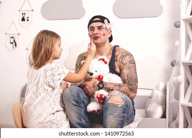Funny time Tattoed father in a cap and his child are playing at home. Cute girl is doing makeup to her dad in her bedroom. Family holiday togetherness