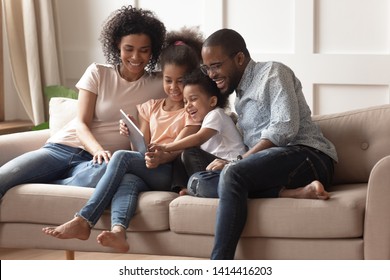 Happy african family having fun with device at home, black parents and little children using digital tablet looking at screen sit on sofa together, cute kids hold computer laugh watch cartoons online