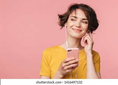 Billede af en glad ung smuk kvinde poserer isoleret over lyserød vægbaggrund og lytter til musik med høretelefoner ved hjælp af mobiltelefon.