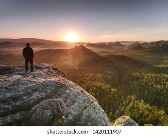 El hombre en el imperio de la roca con las manos en el bolsillo de los pantalones vigila el valle brumoso de la mañana al sol naciente en el horizonte.