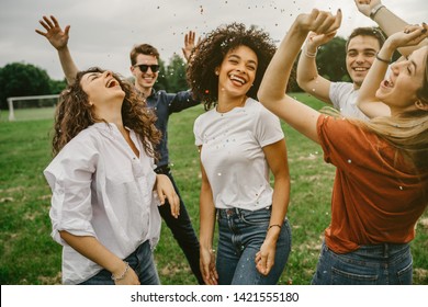 Gruppe von fünf Freunden, die sich im Park amüsieren - Millennials tanzen auf einer Wiese zwischen Konfetti, die in die Luft geworfen werden - Tag der Freiheit und Sorglosigkeit