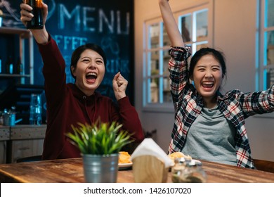 zwei junge hübsche asiatische Mädchen Fußballfan mit Flasche Bier in den Händen Fußball in der Sportbar in der Nacht beobachten. fröhliche Frauen erhoben die Arme und feierten das Siegesfußballspiel im Pub Japan