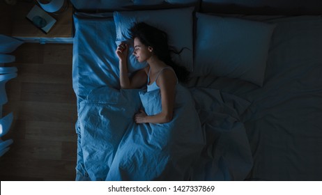 Bovenaanzicht van mooie jonge vrouw die 's nachts gezellig op een bed in zijn slaapkamer slaapt. Blauwe nachtelijke kleuren met koud zwak lantaarnpaallicht dat door het raam schijnt.